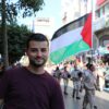 Un jeune homme Khaled Abu-Qare se tient au premier plan et derrière lui se trouve une marhe de scouts. Un homme tient un drapeau palestinien. Palestine, 2021. Crédit: Khaled Abu-Qare