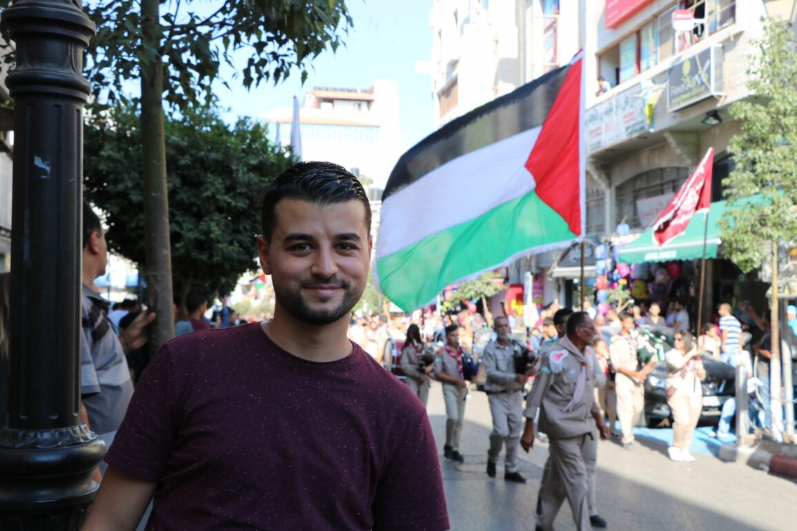 Un jeune homme Khaled Abu-Qare se tient au premier plan et derrière lui se trouve une marhe de scouts. Un homme tient un drapeau palestinien. Palestine, 2021. Crédit: Khaled Abu-Qare