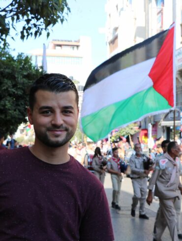 Un jeune homme Khaled Abu-Qare se tient au premier plan et derrière lui se trouve une marhe de scouts. Un homme tient un drapeau palestinien. Palestine, 2021. Crédit: Khaled Abu-Qare